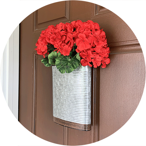 A galvanized metal container with red artificial geraniums hanging on a front door.